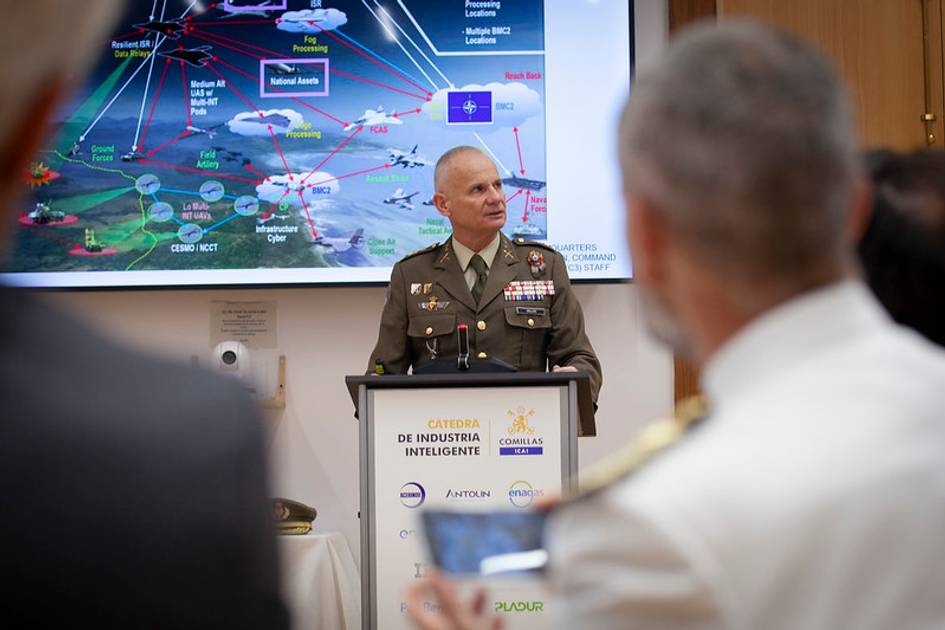 A military officer is presenting a detailed strategic map to an audience of professionals at a conference.