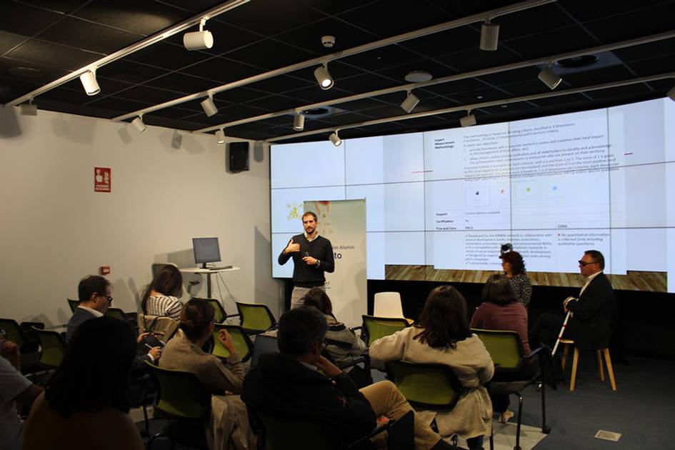 A man is presenting in front of an audience in a seminar room with informational posters on the wall.