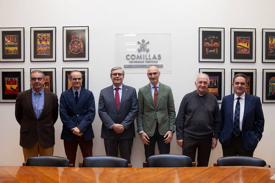 Seis hombres de pie en una sala con el logotipo de la Universidad Comillas y cuadros coloridos en la pared.