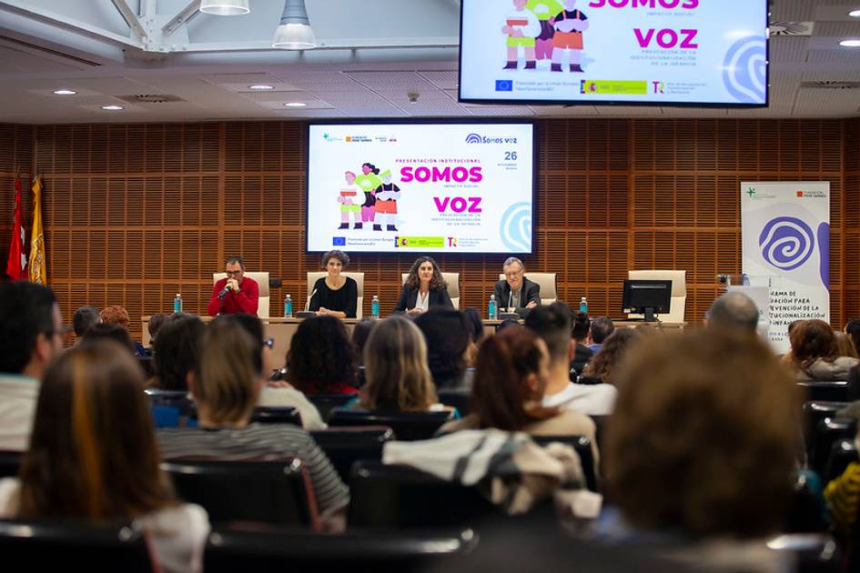 Tres personas están sentadas en un panel bajo un gran cartel que dice 'SOMOS VOZ' en una sala con público.