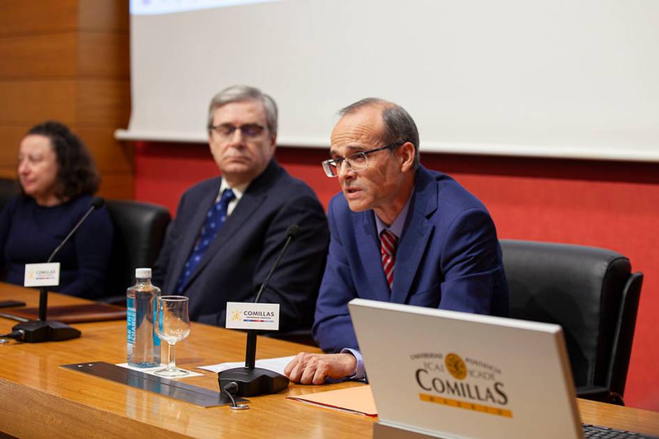 Un hombre habla desde un podio en un evento en la universidad Comillas, acompañado por otros dos participantes.