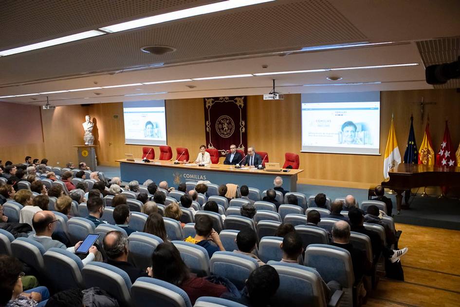 Personas asistiendo a una conferencia en un auditorio con paneles de presentación iluminados al fondo.