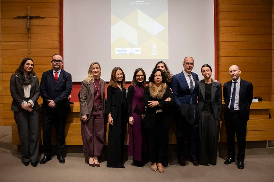 Grupo de ocho personas posando juntas en una sala con sillas y un crucifijo en la pared.