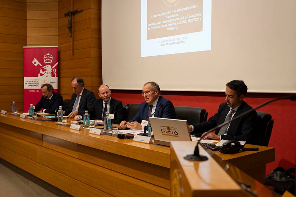 Cinco hombres participan en una conferencia en una sala con audiencia y un proyector encendido mostrando una presentación.