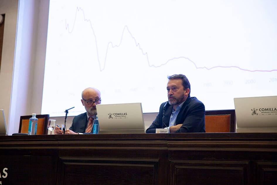 Dos hombres en un panel de discusión sentados frente a micrófonos en un salón con gráficos de fondo.