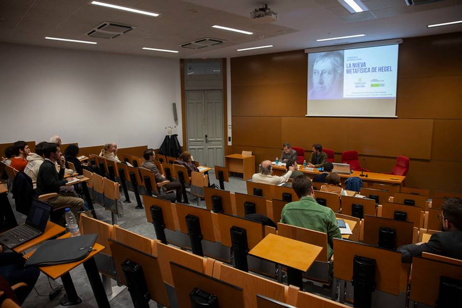 Un grupo de personas asiste a una charla sobre la Nueva Metafísica de Hegel en un aula con sillas dispuestas en filas.