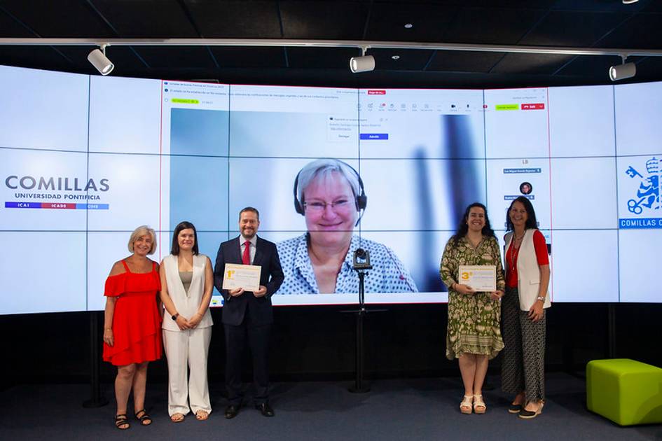 Una ceremonia de entrega de diplomas con varios participantes en un aula moderna y una persona conectada a través de videoconferencia.