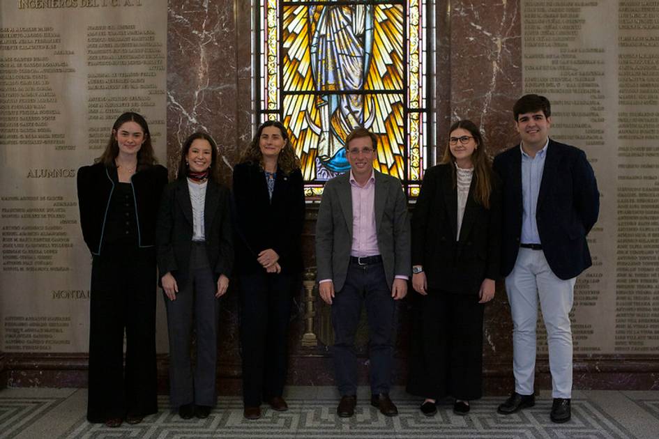 Grupo de seis personas posando frente a una ventana de vitral en un entorno académico.