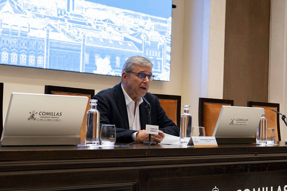 Un hombre habla en una conferencia mientras está sentado ante un atril con el logo de Comillas en un ambiente académico.