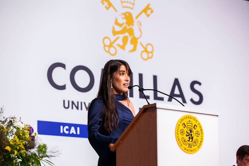 A woman speaking at a podium with the Comillas University logo in the background.