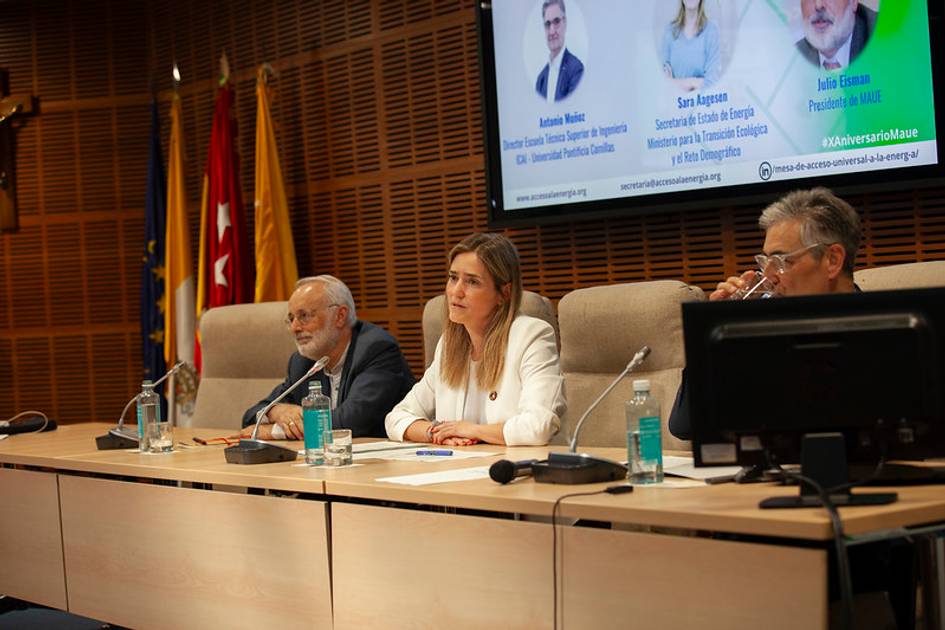 A panel discussion at a conference with four panelists, including three men and one woman, seated at a table with microphones and water bottles, in front of a presentation screen displaying names and titles.