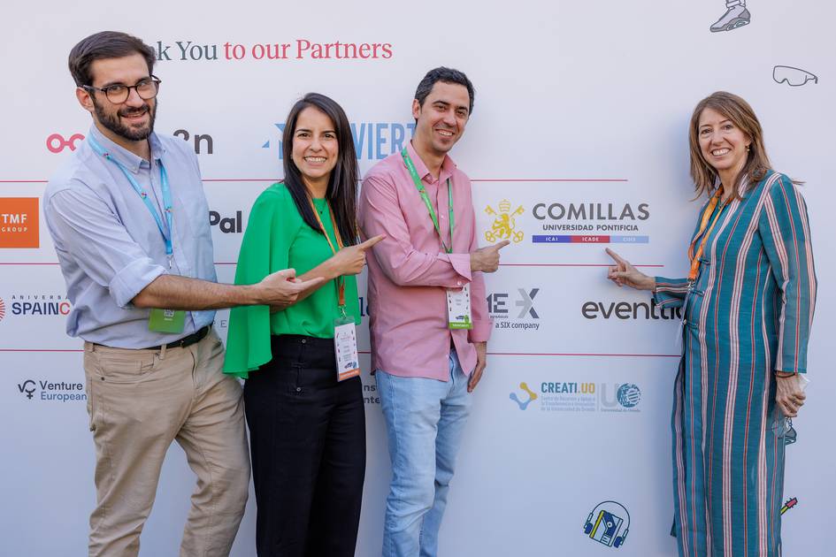 Cuatro personas están posando y señalando un logotipo de la Universidad Pontificia Comillas en un evento.
