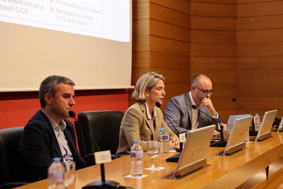 Tres personas sentadas en una mesa durante una conferencia con pantallas al fondo.