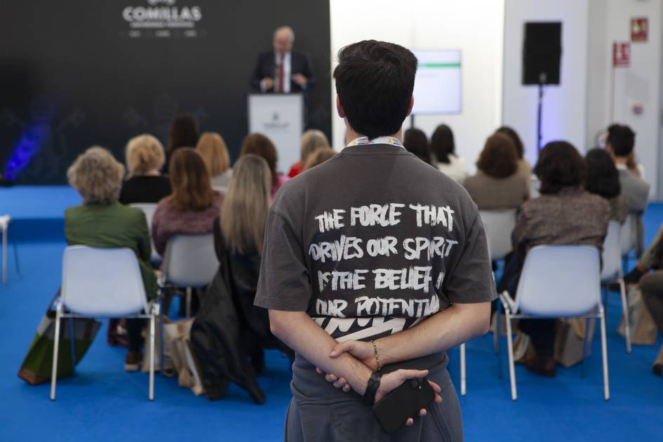 Un grupo de personas asiste a una conferencia mientras un hombre con una camiseta con mensaje observa desde atrás.