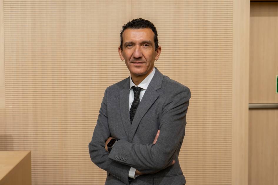 Un hombre de pie con brazos cruzados, vestido con un traje gris y una corbata negra, frente a un fondo de madera claro.