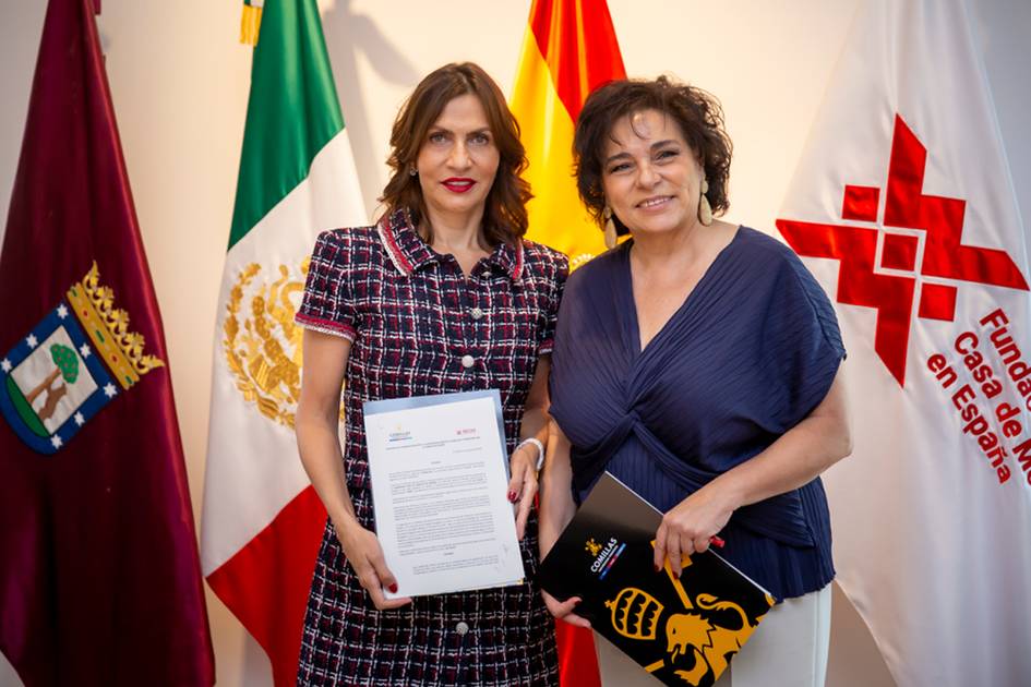 Two women stand smiling, holding a document together, with flags of Mexico, Spain, and the Red Cross in the background.