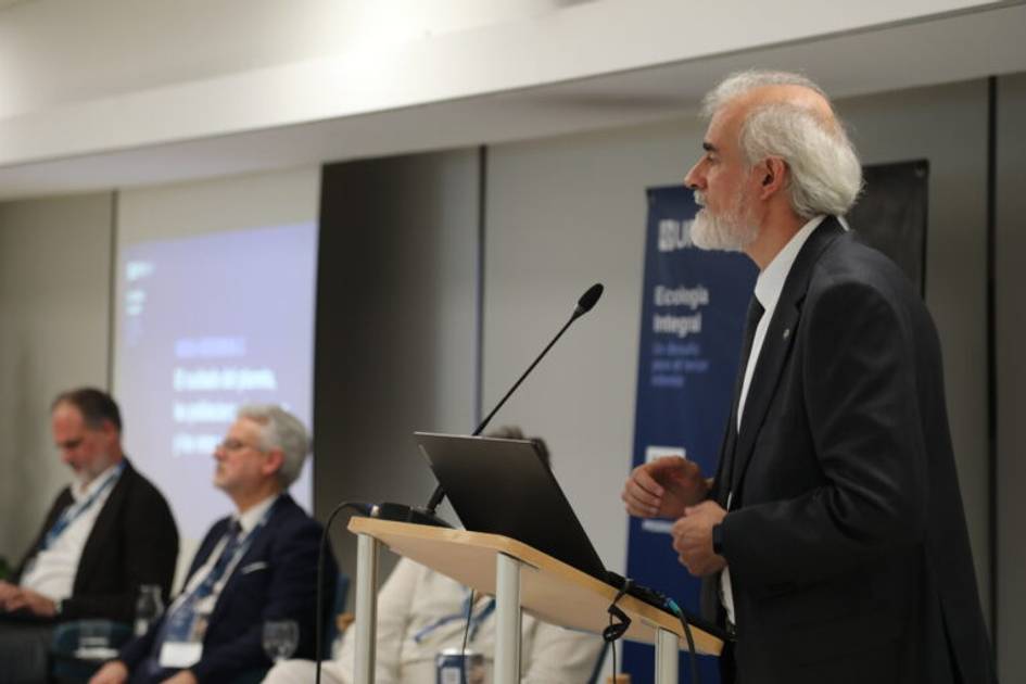 A man with white hair speaking at a podium during a panel discussion with three other participants seated in the background.