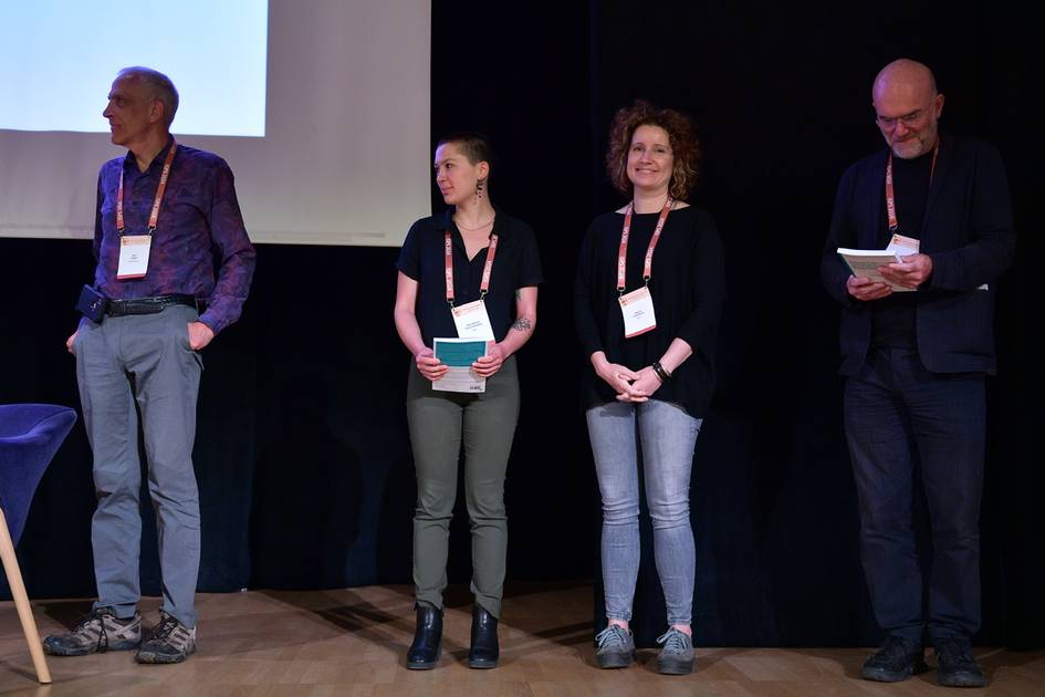 Four people standing on a stage at a conference, wearing name tags and looking towards the audience.