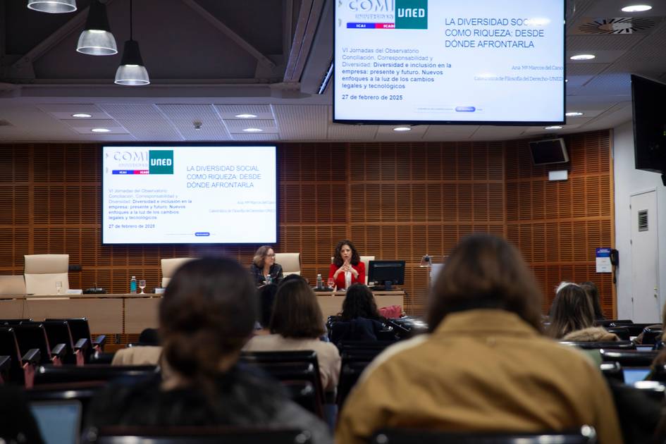 La imagen muestra un evento con ponentes en un escenario y una audiencia atenta en un auditorio.