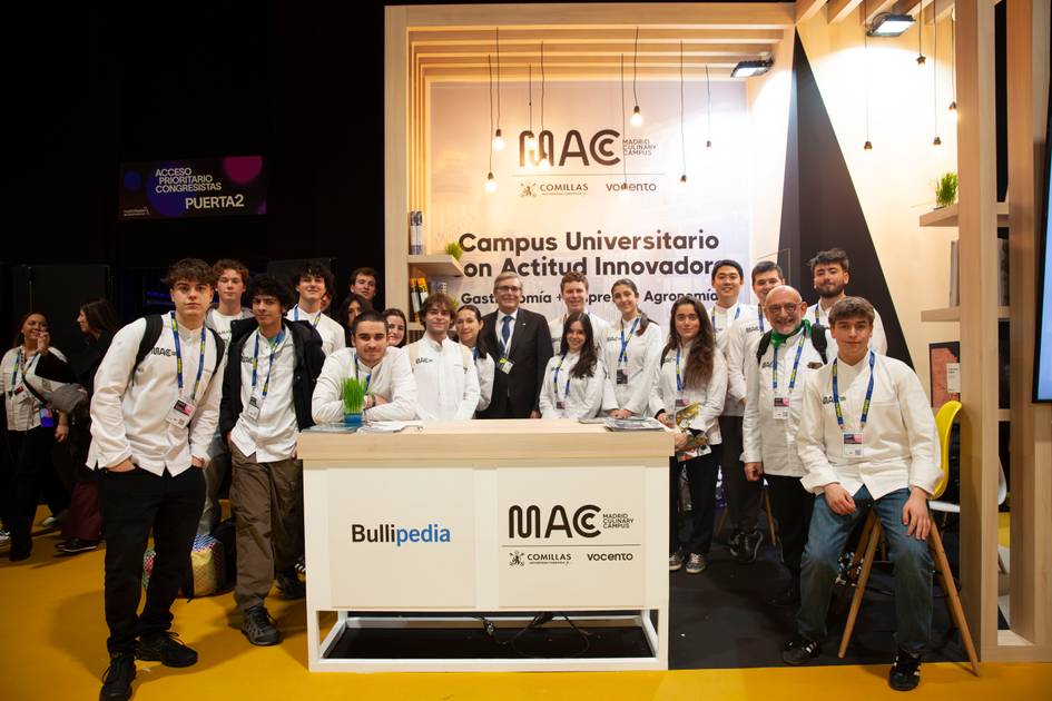 Un grupo de jóvenes en batas blancas posa sonriente detrás de un mostrador en un evento, rodeados de carteles promocionales.