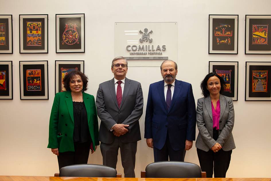 Four individuals are standing in a formal office setting in front of a wall with a 'Comillas Universidad Pontificia' sign and decorated with colorful art pieces.