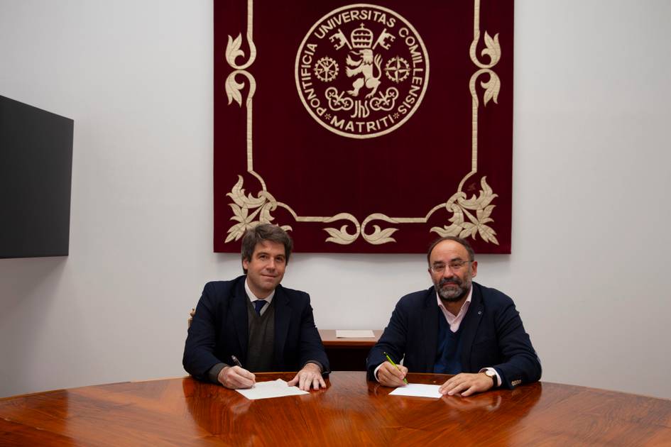 Two men in suits sitting at a table signing documents with a red university banner in the background.