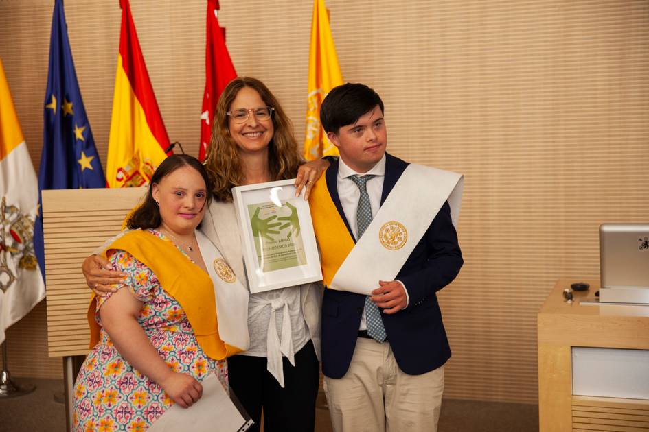A woman and two young graduates holding certificates pose together in a room decorated with flags.
