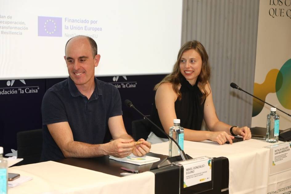 Dos personas sentadas en una mesa durante un evento, una de ellas sonriente y la otra mirando hacia adelante.