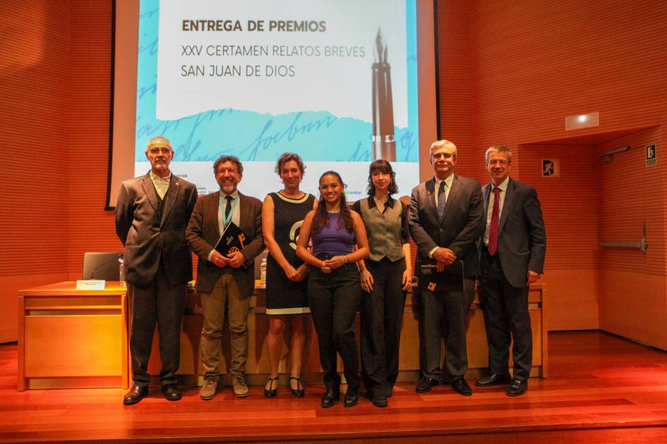 Group of seven people standing on stage at an awards ceremony for a short story contest named 'XXV Certamen Relatos Breves San Juan de Dios'.