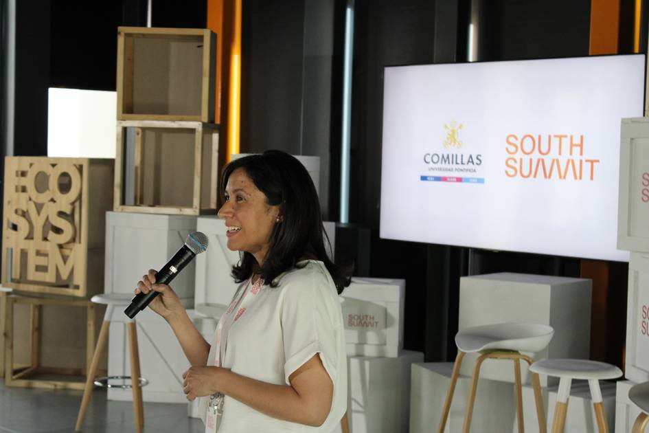 A woman is speaking into a microphone at a conference with logos displayed in the background, including 'Comillas' and 'South Summit.'