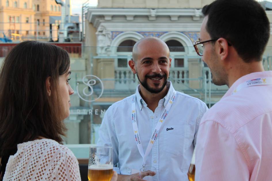 Three people having a conversation at a social event, with one man smiling and holding a beer, surrounded by two others.