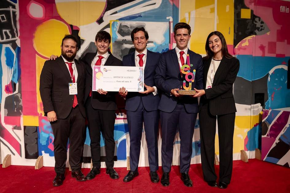 Un grupo de cinco personas posando sonrientes en un evento, sosteniendo un premio y un cheque frente a un fondo colorido.