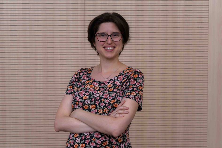 Una mujer sonriente con gafas y una blusa de flores posa con los brazos cruzados frente a una pared con persianas venecianas.