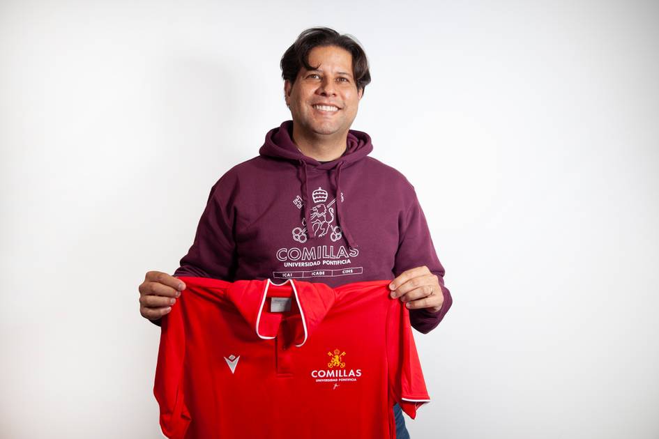 A man in a purple hoodie smiles while holding a red Comillas University sports jersey.