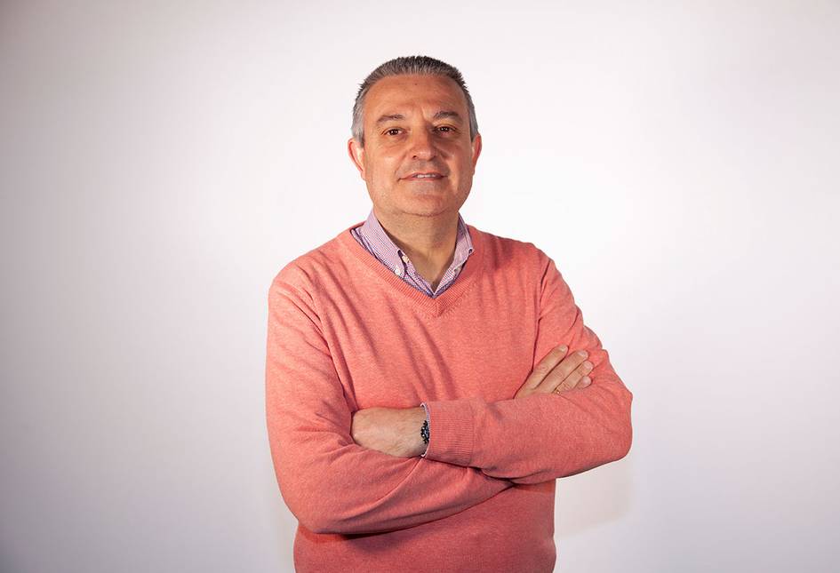 A man in a pink sweater and blue shirt standing with crossed arms against a white background.