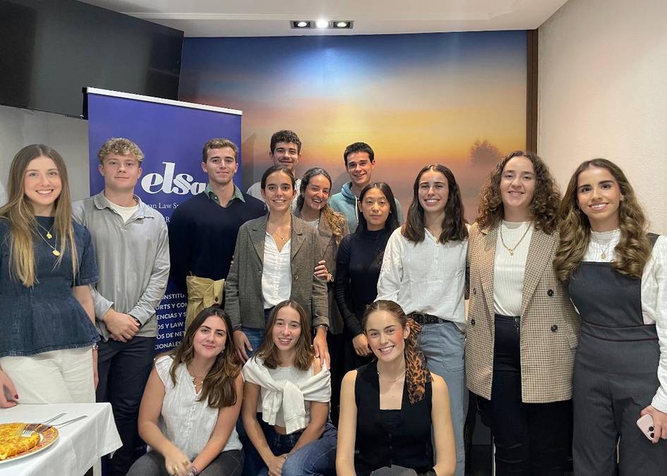 Grupo de jóvenes sonriendo para la cámara en un evento o reunión en una sala con un cartel de 'elsa' de fondo.