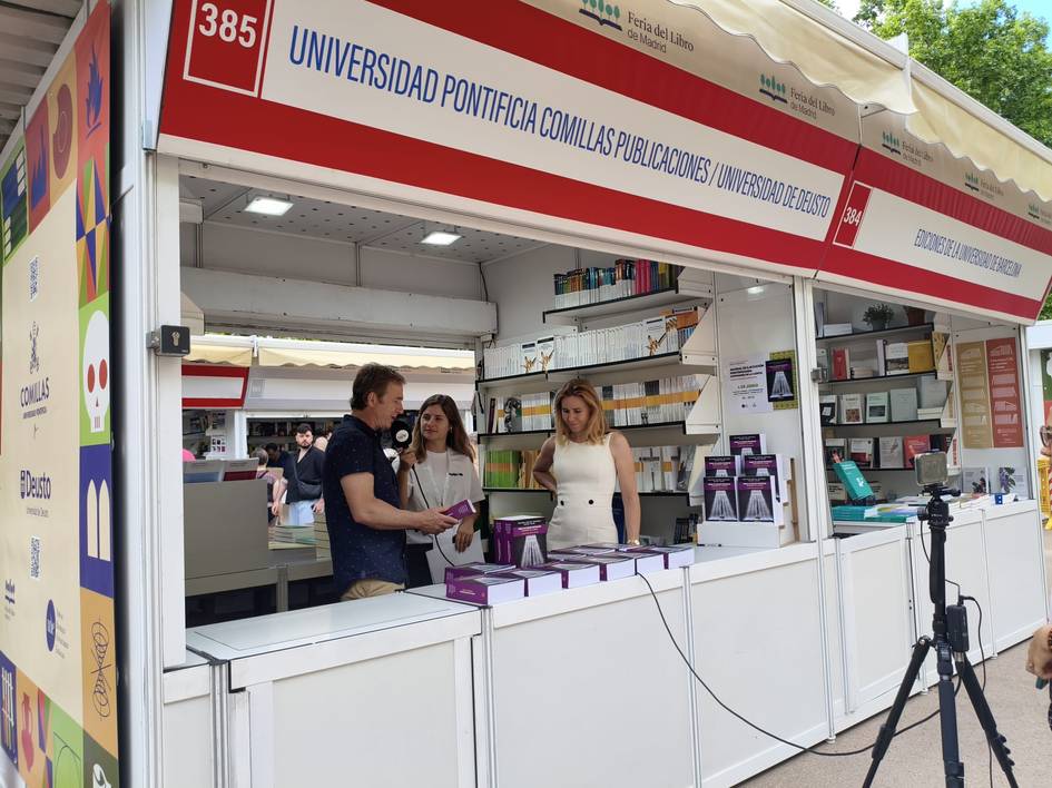Una feria del libro con un stand de la Universidad Pontificia Comillas donde hay personas interactuando.