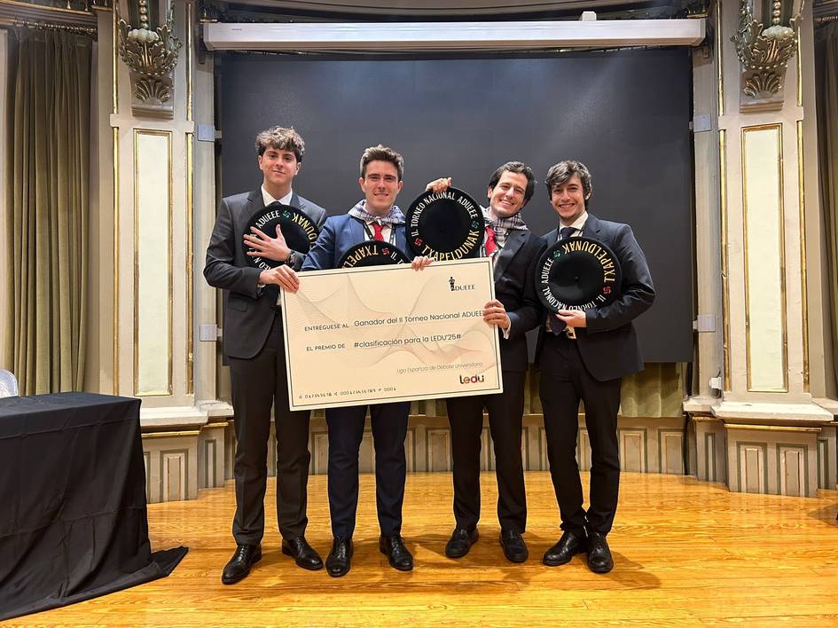 Four young men in suits holding awards and a large check in a ceremonial hall.