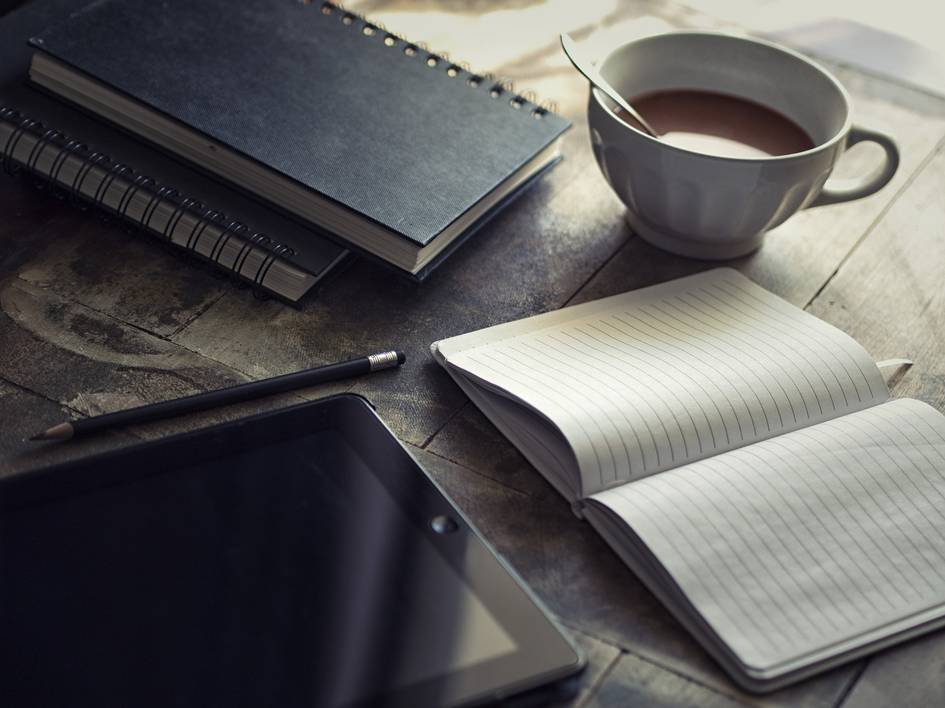 A cozy workspace with a cup of tea, notebooks, a pencil, and a tablet on a wooden table.