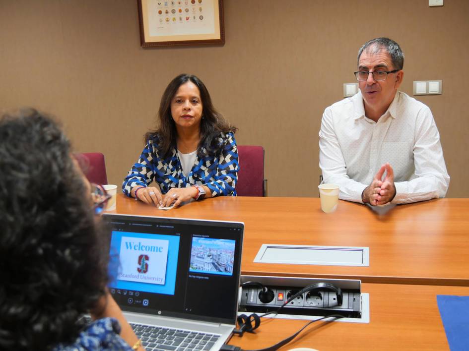 La imagen muestra a dos personas sentadas en una mesa durante una reunión, con una computadora portátil y una presentación visible en la pantalla.
