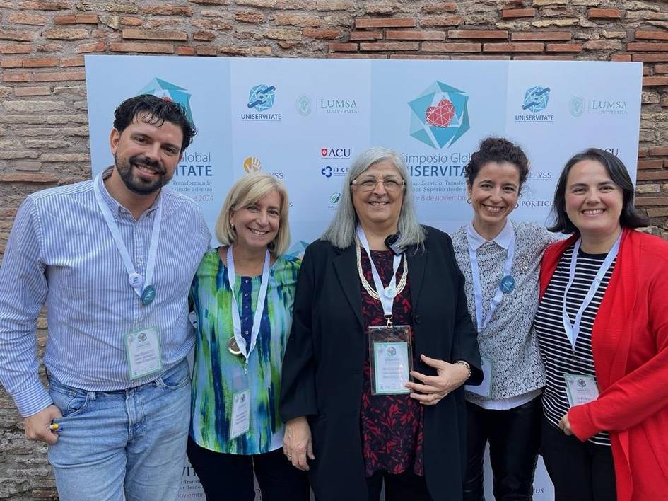 Cinco personas sonriendo para la foto durante un evento académico con banners de universidades de fondo.