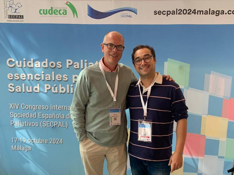 Two men smiling at a conference with a banner about public health and palliative care in the background.