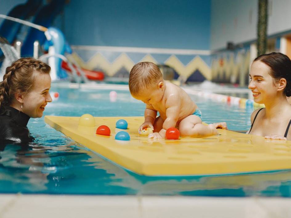 A baby plays with colorful balls on a floating mat in a swimming pool, flanked by two smiling women.