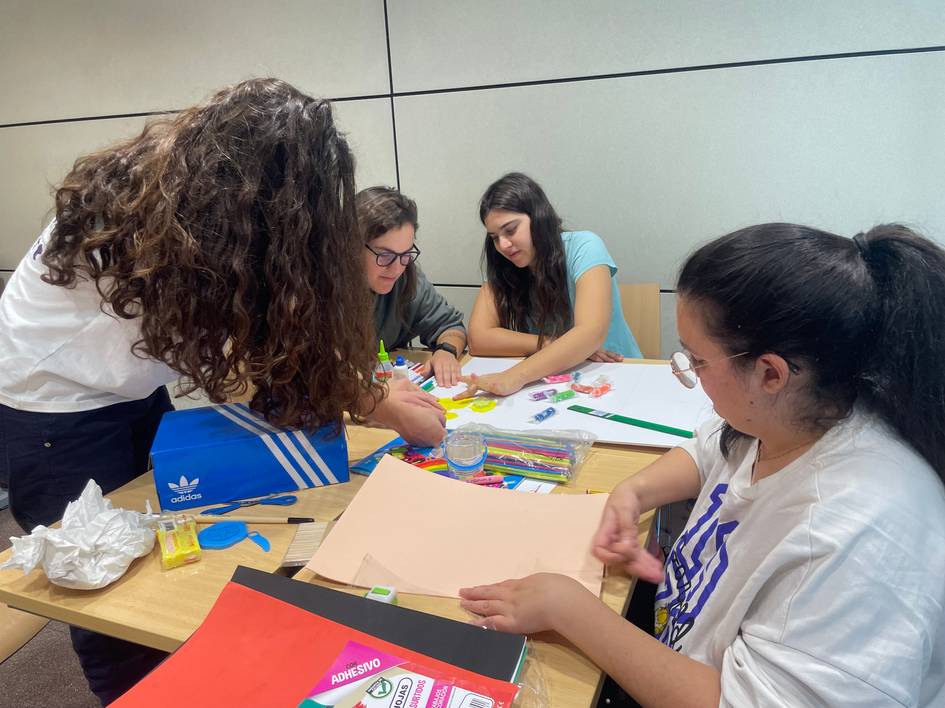 Un grupo de jóvenes se encuentra sentado alrededor de una mesa realizando manualidades con papeles de colores y otros materiales.