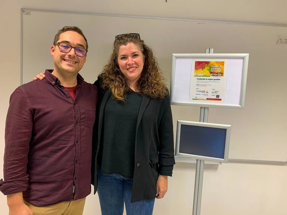 Un hombre y una mujer sonriendo, posando juntos en un espacio de oficina con un cartel al fondo.