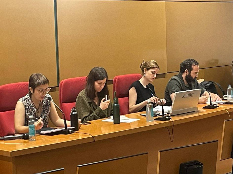 Four people sitting at a conference table, each using a laptop or taking notes, in a meeting room with red seats.