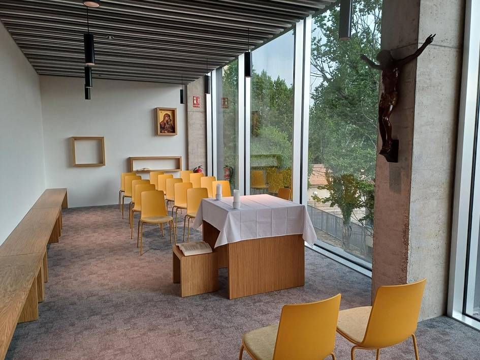 La imagen muestra un interior de una sala con bancos, mesas y un altar junto a grandes ventanales que permiten la entrada de luz natural.