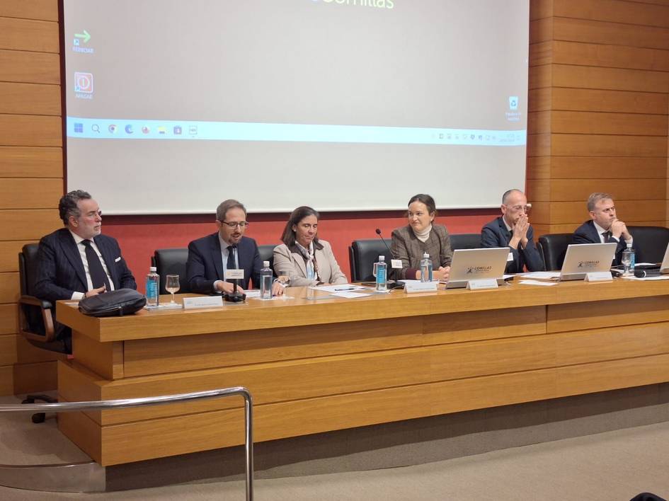 Cinco personas están sentadas en una mesa durante una conferencia en una sala con una pantalla grande al fondo.