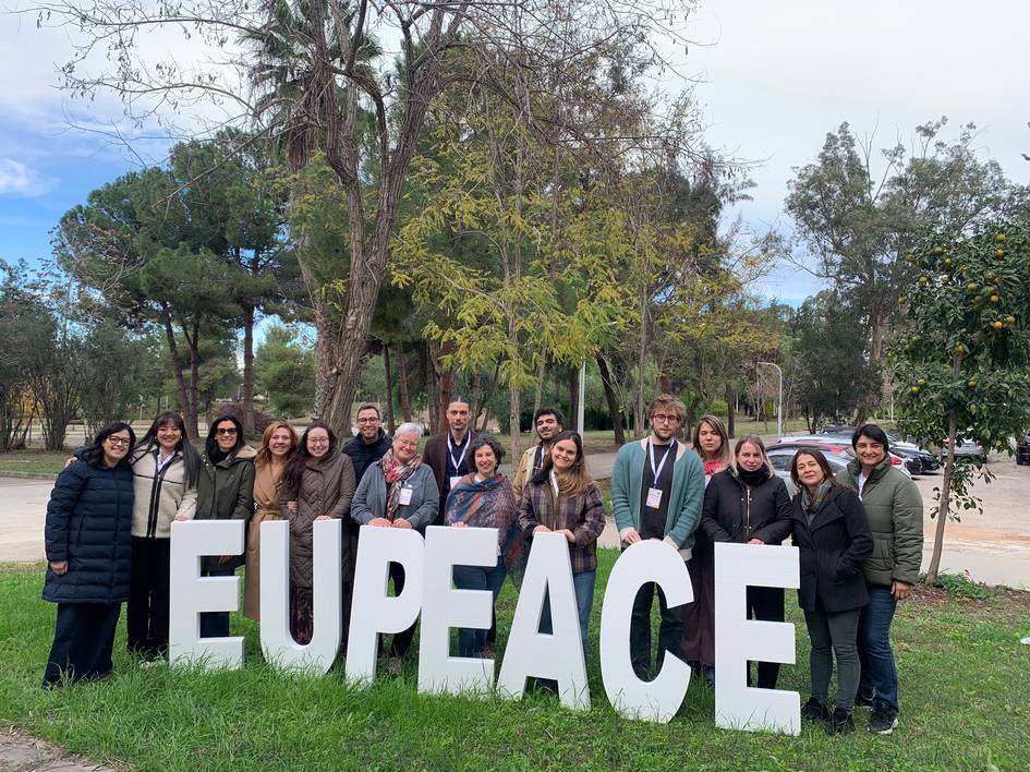 Un grupo de personas posa frente a un cartel grande que dice 'EUPEACE' en un entorno natural.