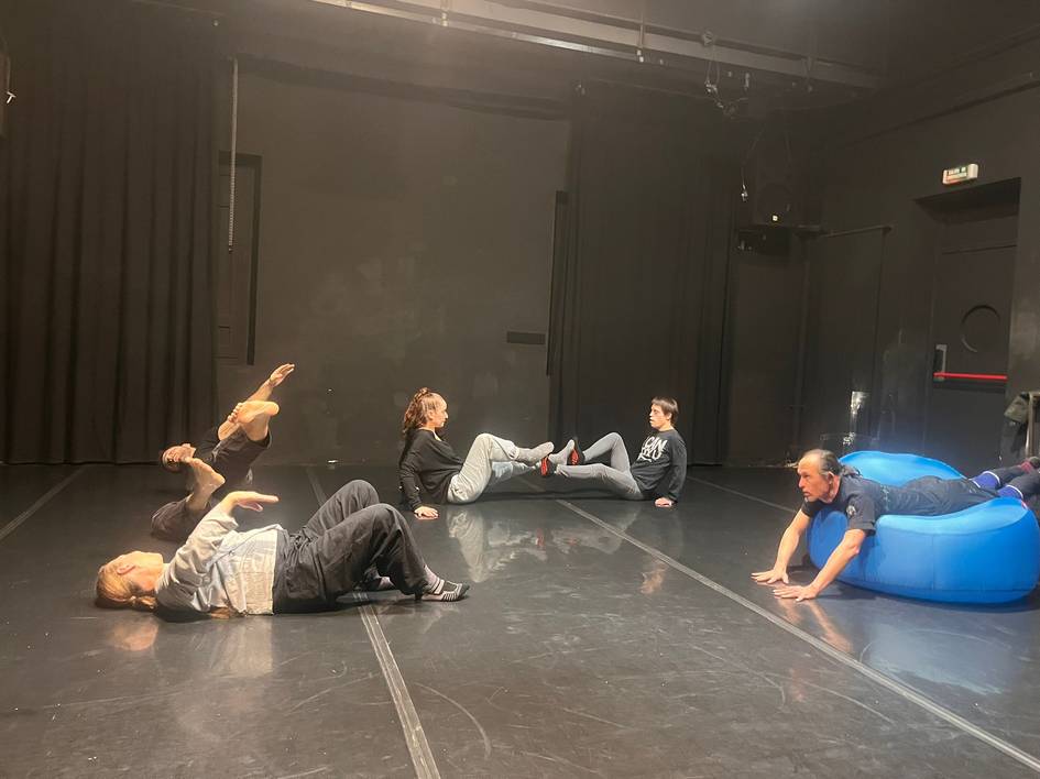 Un grupo de personas descansa en un escenario oscuro, realizando diferentes posturas sobre el suelo y un sofá inflable.